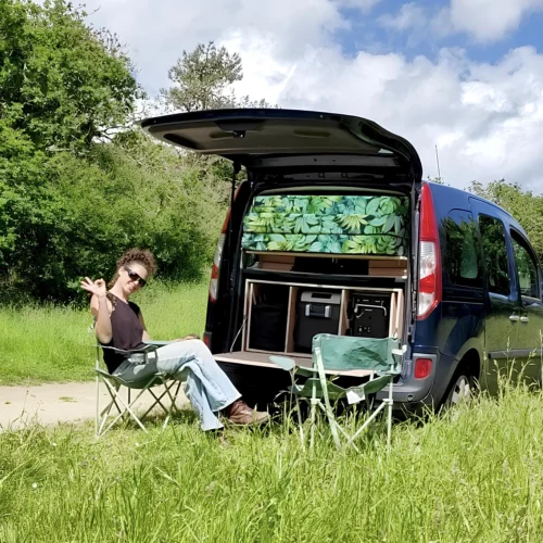 Ma malle d'enfer - Transformer votre véhicule de tout les jours en van aménagé avec nos kits d'aménagement - femme qui boit le café, asssise sur son matelas, regardant dehors par la porte arrière de sa voiture qui a été transformée en van aménagé grâce à Ma Malle d'Enfer