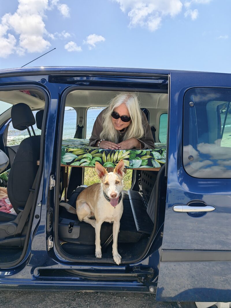 Ma malle d'enfer - Transformer votre véhicule de tout les jours en van aménagé avec nos kits d'aménagement - femme qui boit le café, asssise sur son matelas, regardant dehors par la porte arrière de sa voiture qui a été transformée en van aménagé grâce à Ma Malle d'Enfer